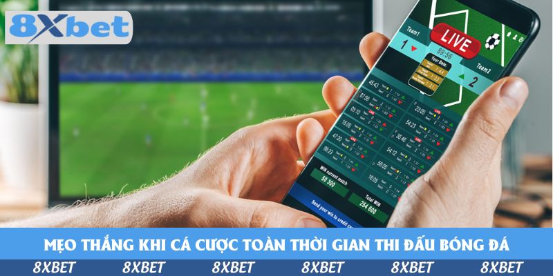 Người cầm điện thoại xem bảng cược trực tiếp trước màn hình đang phát trận bóng đá