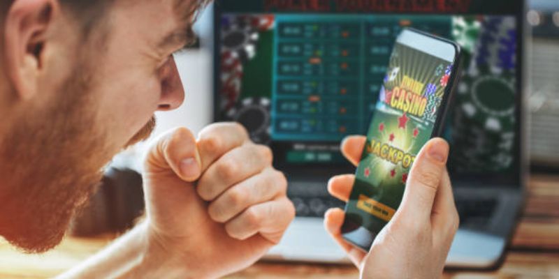 Người đàn ông nắm chặt tay hào hứng nhìn điện thoại hiện chữ Casino Jackpot khi chơi 8xbet game, phía sau là laptop mở bảng cược